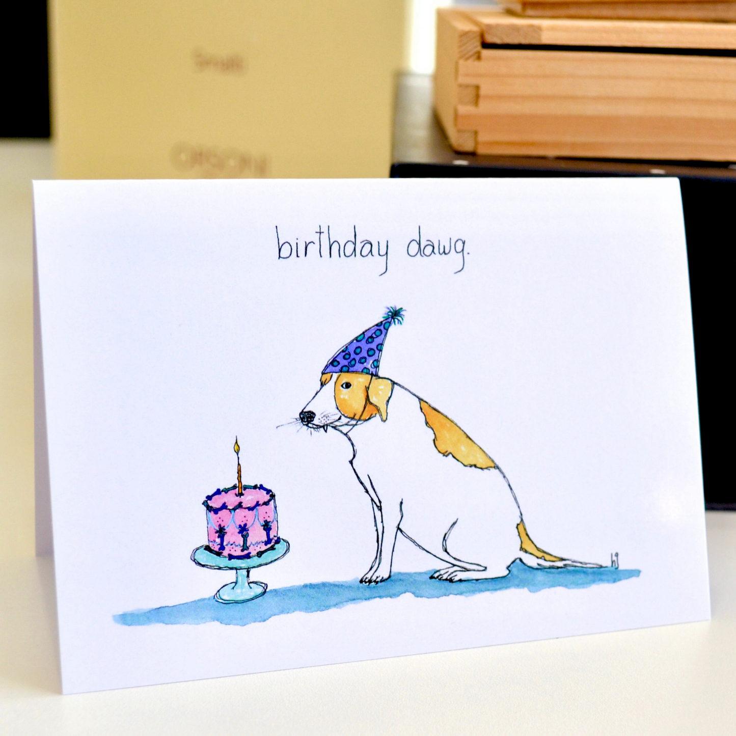 A lovely old tan and white dog with a snaggle tooth sitting next to a decorated birthday cake topped with a candle.