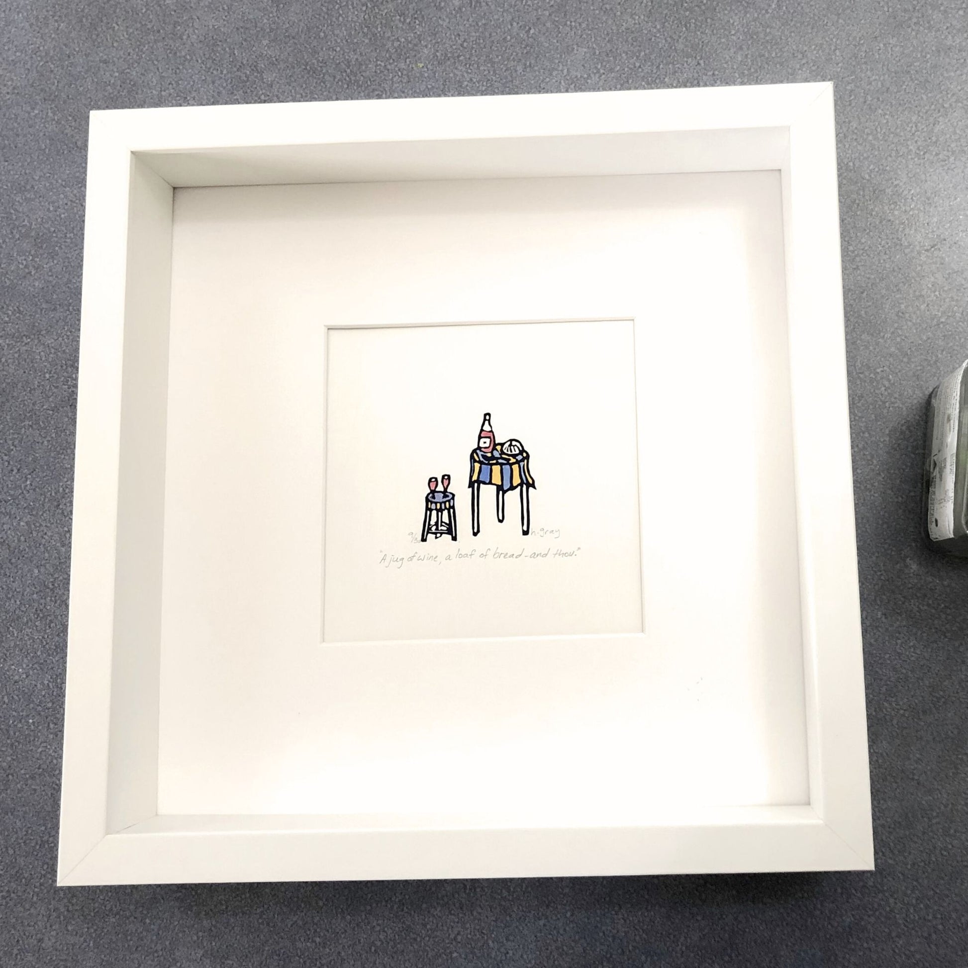 A lino print of a bottle of wine, glasses and bread on a small round table with a blue and yellow striped tablecloth. Hand-coloured with watercolour paint.