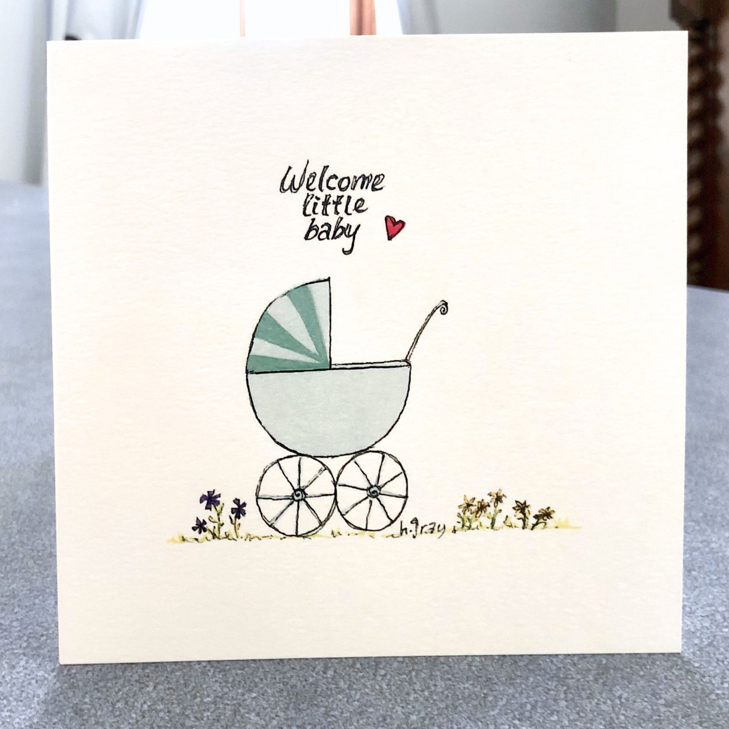 Square baby card, with an old fashioned green pram with some flowers on the ground.