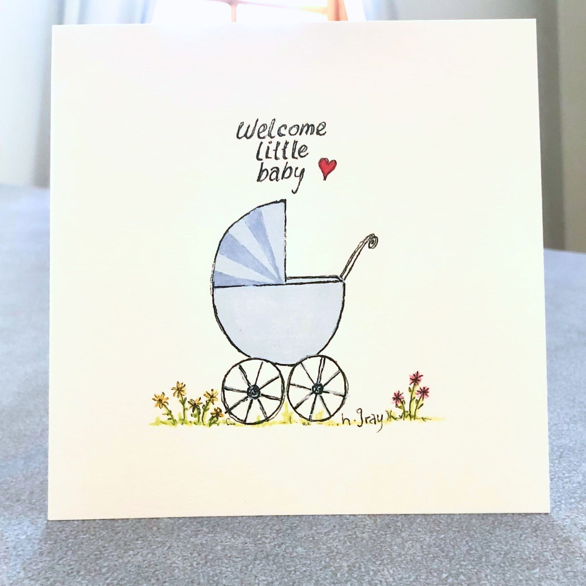 Square baby card, with an old fashioned blue pram with some flowers on the ground.