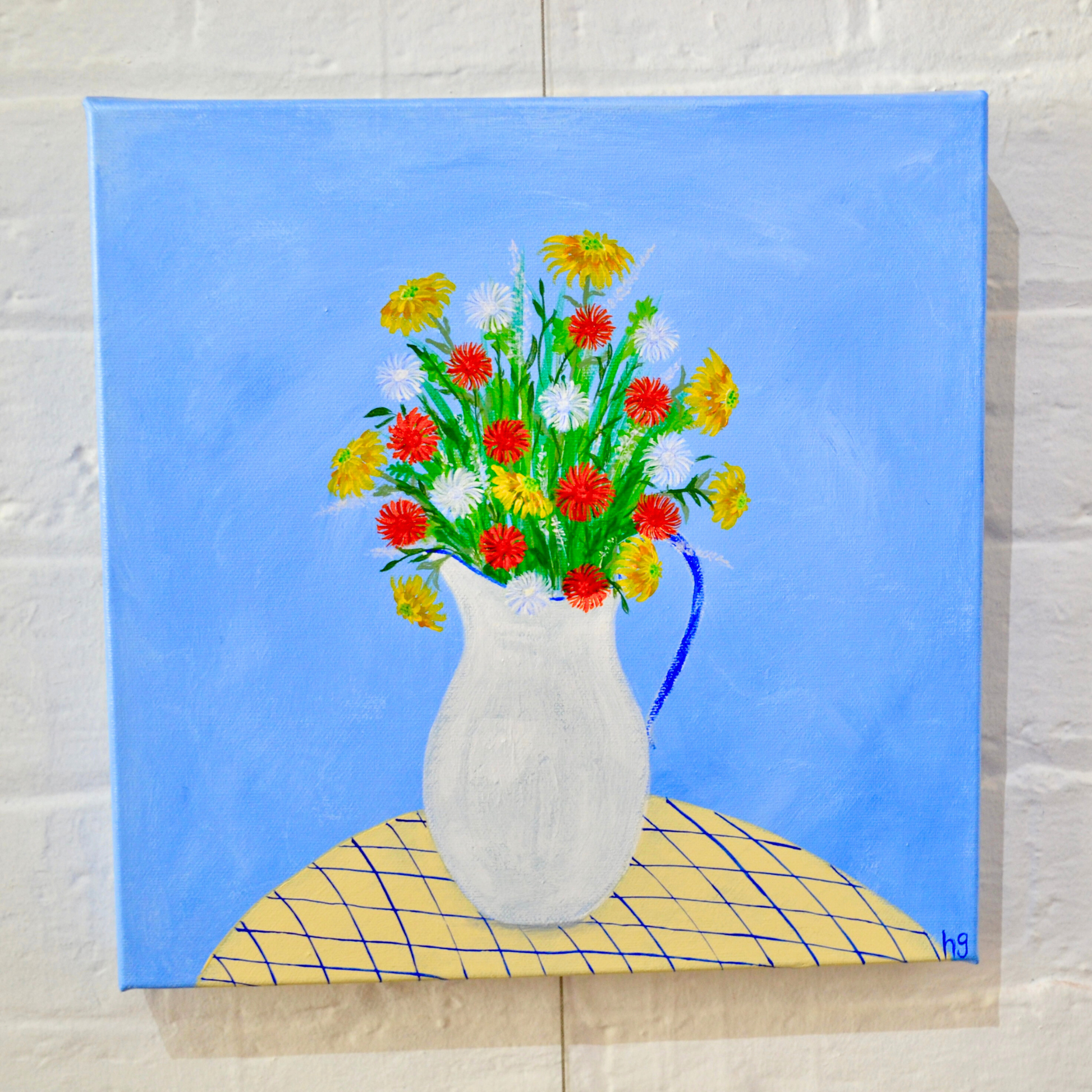 A floral still life in a white and blue-rimmed enamel jug, sitting on a yellow patterned tablecloth, with a blue background. Acrylic paint on canvas, measuring 30x30 centimetres.