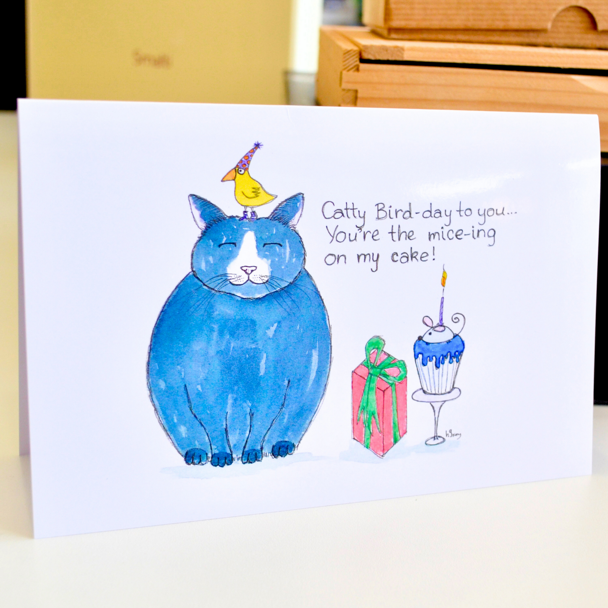 A small yellow bird wearing a party hat sitting on a happy cat's head, with a birthday cake topped by a mouse candle, with a present. The title reads 'Catty Bird-Day to you...you're the mice-ing on my cake!".