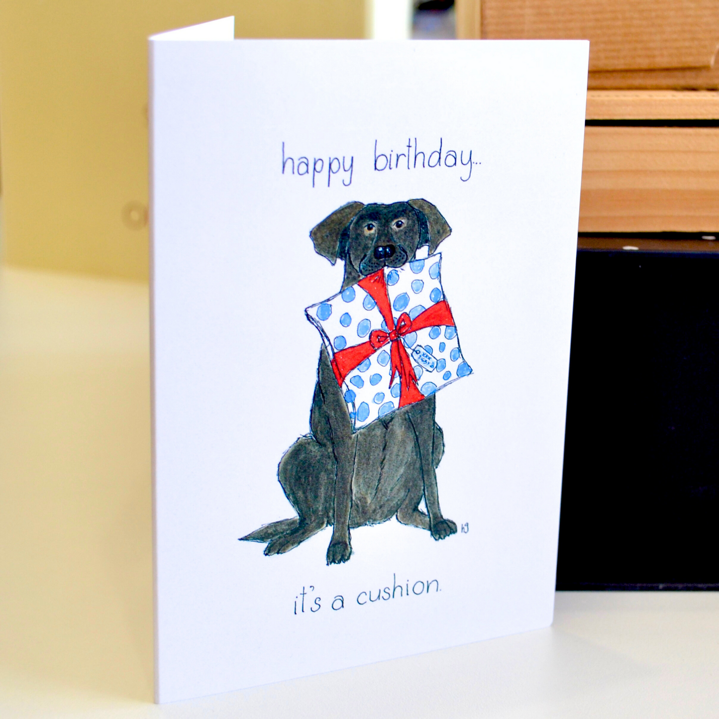 A chocolate labrador carrying in his mouth a cushion wrapped as a gift. The text reads "Happy birthday...it's a cushion.".