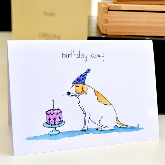 A lovely old tan and white dog with a snaggle tooth sitting next to a decorated birthday cake topped with a candle. 