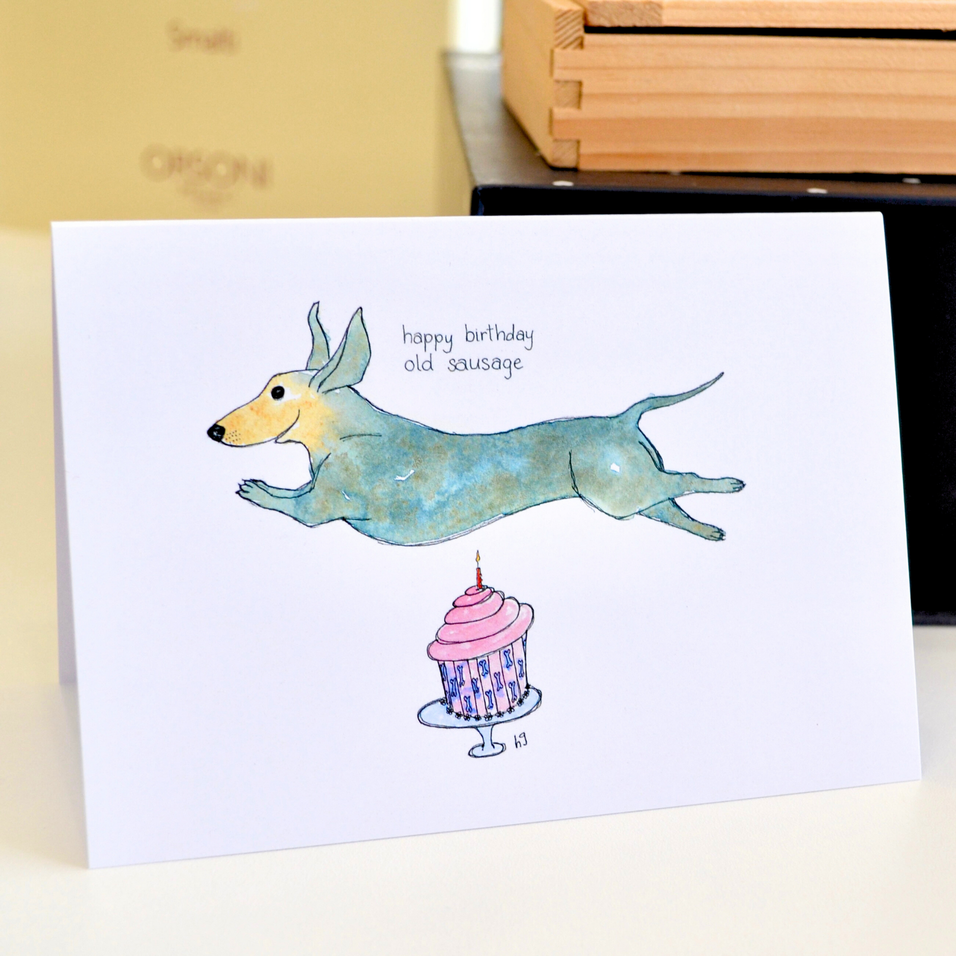 A sausage dog joyfully leaping over a pink birthday cupcake with a candle. The caption reads "Happy birthday old sausage".