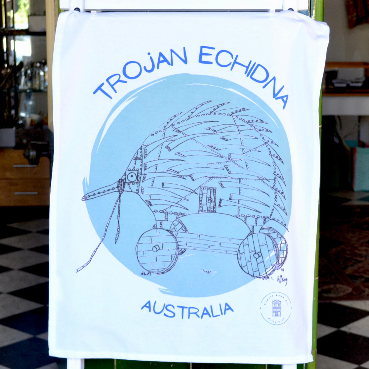 White, 100% soft cotton Tea towel, with a humorous drawing of a Trojan echidna in black ink on a light green circular background, and the words "Trojan Echidna Australia" above and below the echidna in a blue font. Comes with a decorative cardboard band.