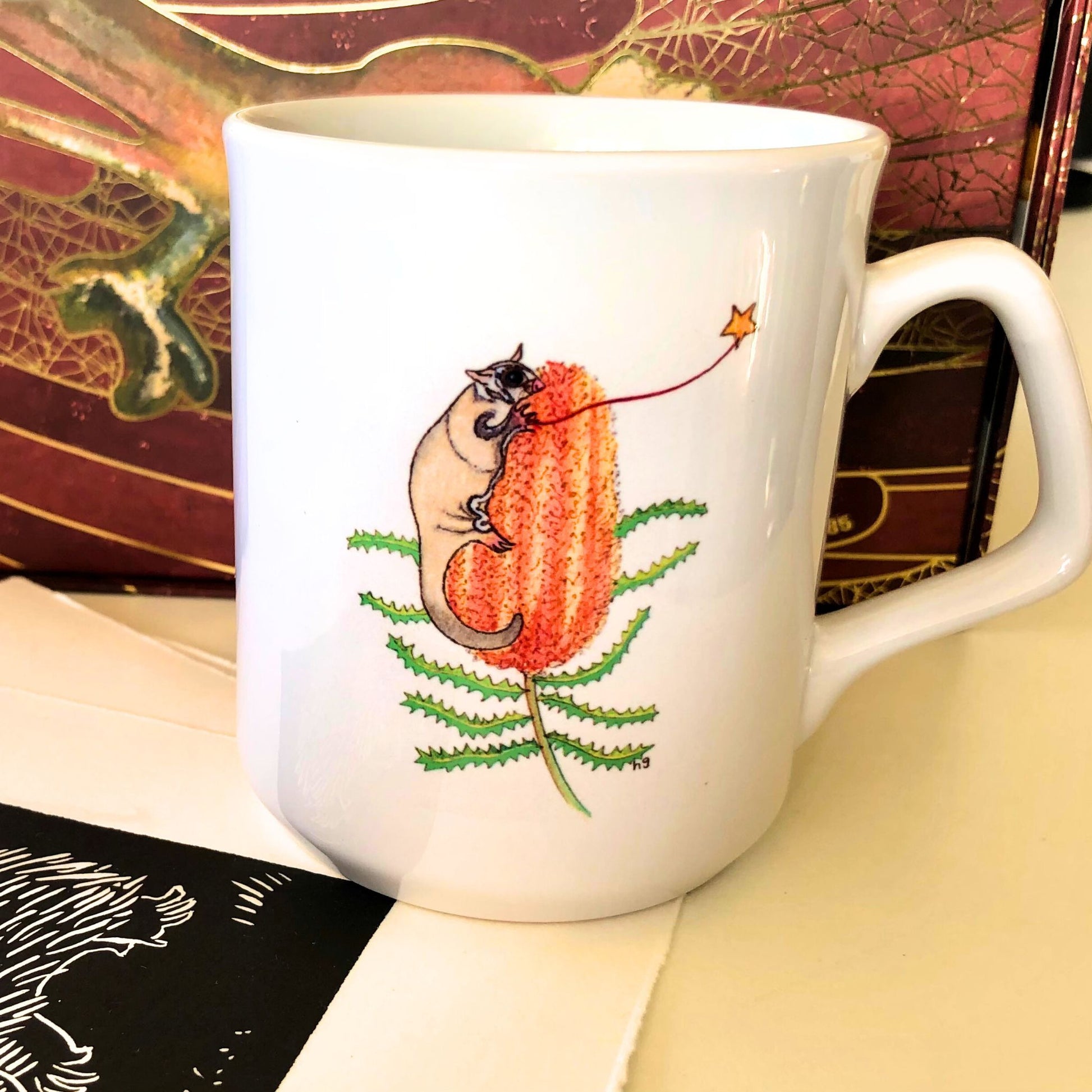 White coffee mug with a sugar glider possum on a banksia plant, holding a gold star on a string. From original artwork.