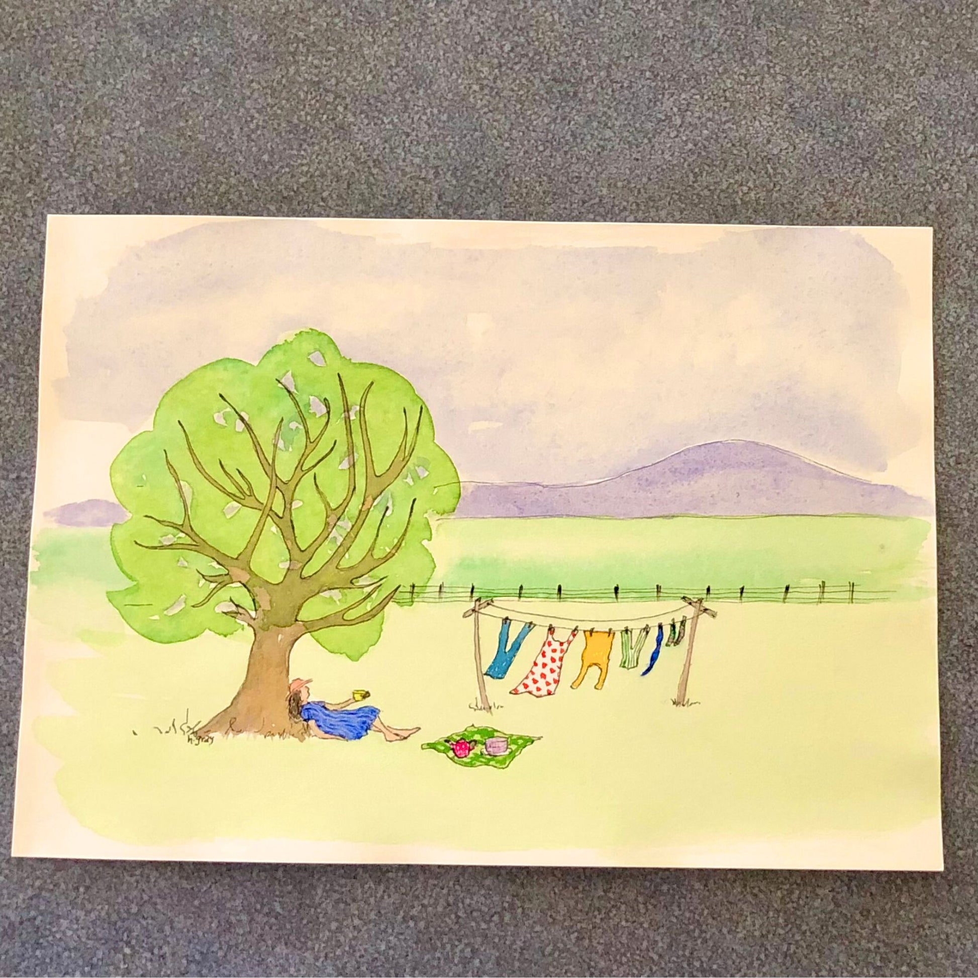 An original watercolour by Helen Gray showing a woman lying under a tree, drinking a cup of tea, and watching clothes dry on a clothesline. There is a small picnic cloth underneath a teapot and cake. There is a mountain in the background and a fence in the foreground.