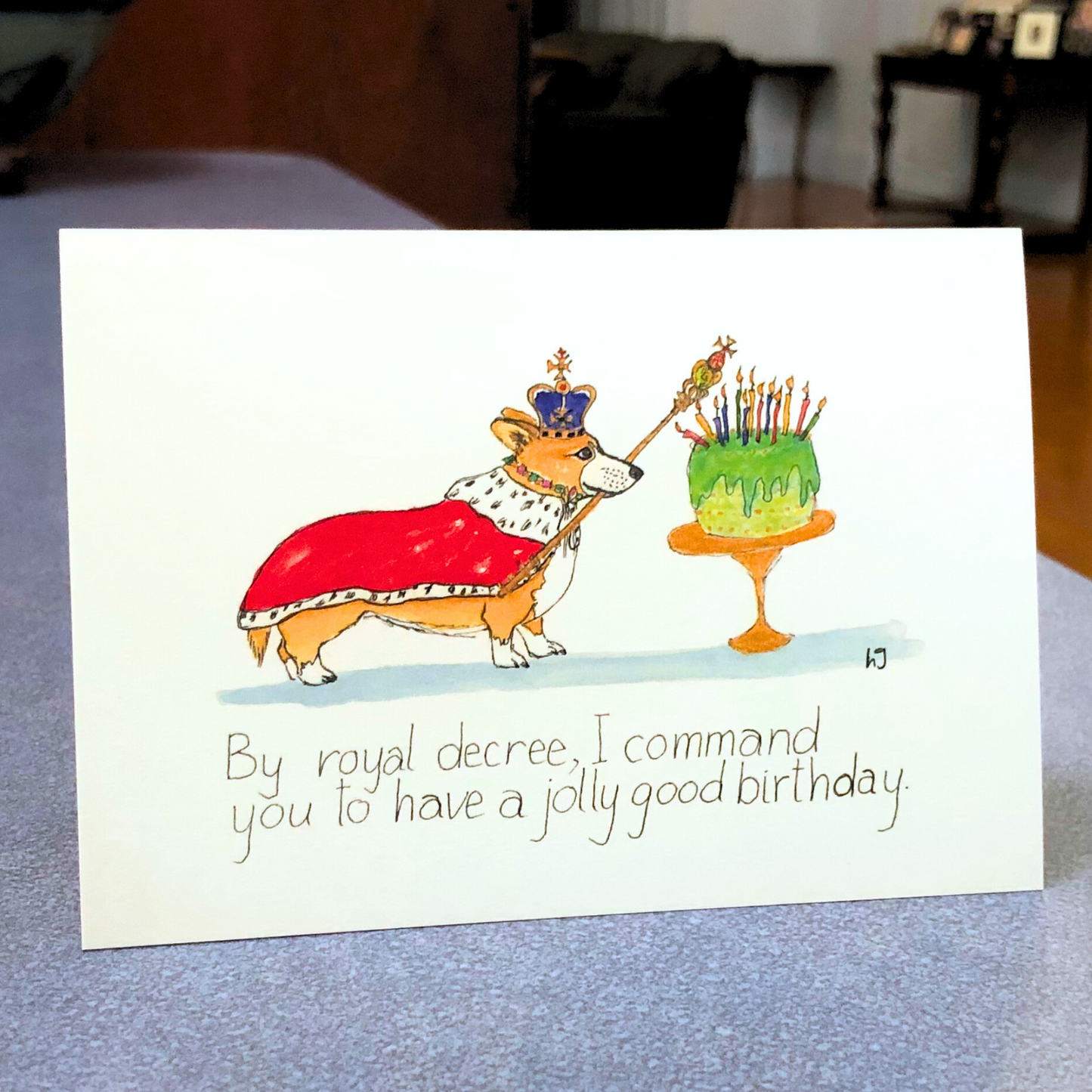 A corgi wearing a red royal cape, with a crown and a sceptre, next to a green birthday cake adorned with many candles. The caption reads " By royal decree, I command you to have a jolly good birthday."